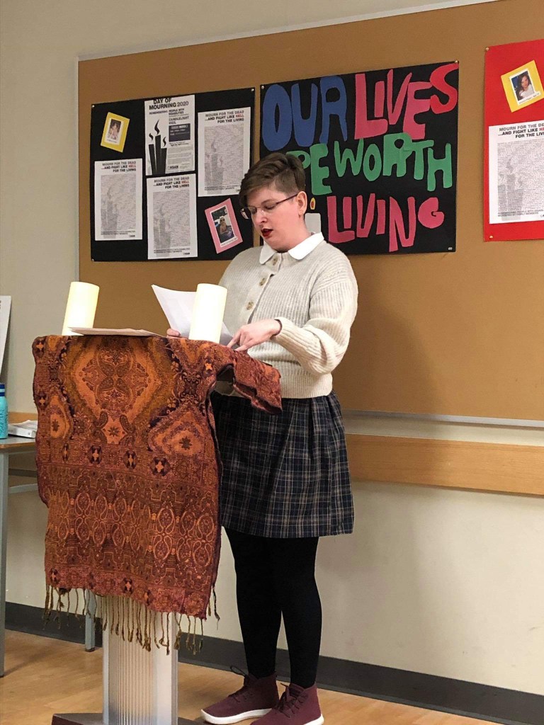 Image Description: Me, a pale blonde woman wearing a plaid dress and cream coloured sweater standing at a podium, reading names off of a list for the Disability Day of Mourning