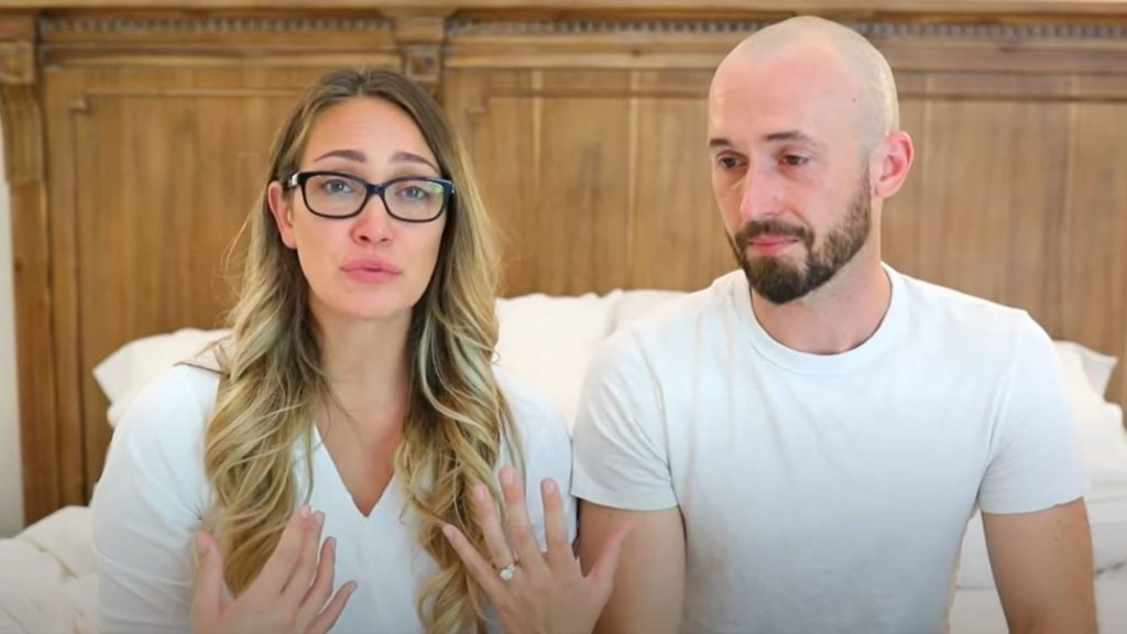 Image description: A white couple Myka and James Stauffer sit on a white bed while both wearing white shirts. Myka has dyed blonde hair and is wearing glasses, James has a beard and is bald