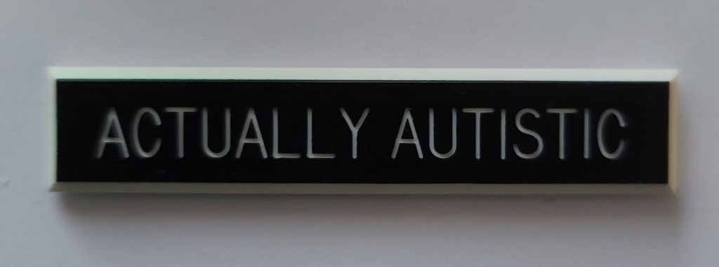 Image Description: Black Label with White writing that says "Actually Autistic"
